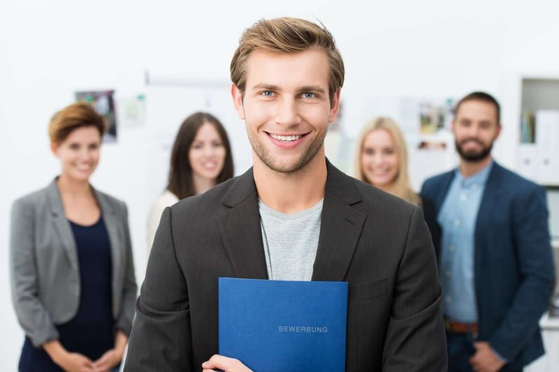 Successful smiling young male job applicant holding a blue file with his curriculum vitae posing in front of his new work colleagues or business team